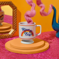 The Brass Monkey Rise & Sigh Pedestal Mug, featuring a rainbow design, sits on round orange platforms with pink wool steam, as a gold-framed mirror and blue teapot add vintage charm to the colorful scene.