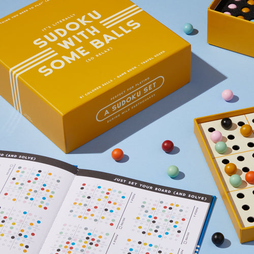 A yellow box labeled Sudoku With Some Balls Sudoku Game Set by Brass Monkey rests on a light blue surface beside a Sudoku board with colored balls and an open setup booklet—a travel-ready game.