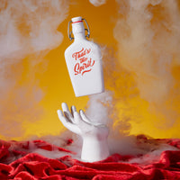 A Brass Monkey Thats The Spirit Porcelain Flask floats above a white hand, amidst swirling smoke, set against a yellow backdrop with red velvet fabric below.