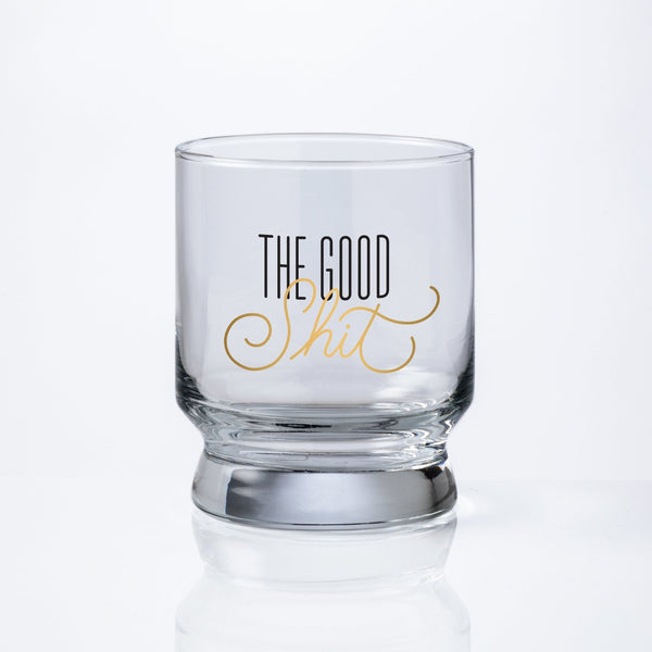 A clear, pedestal-shaped lowball glass by Brass Monkey, featuring THE GOOD Shit in black and gold lettering, shown against a plain white background.
