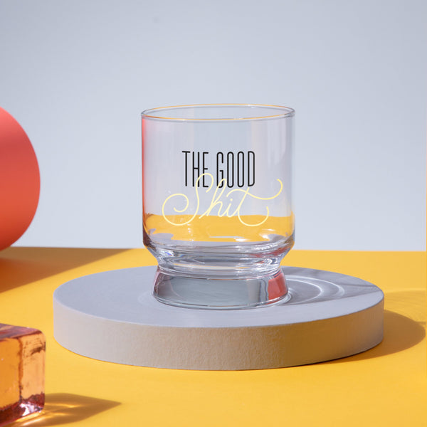 The Good Shit Lowball Glass by Brass Monkey, featuring its signature phrase, rests on a round gray platform against a yellow surface with a pastel backdrop. An orange object and clear orange block sit nearby.
