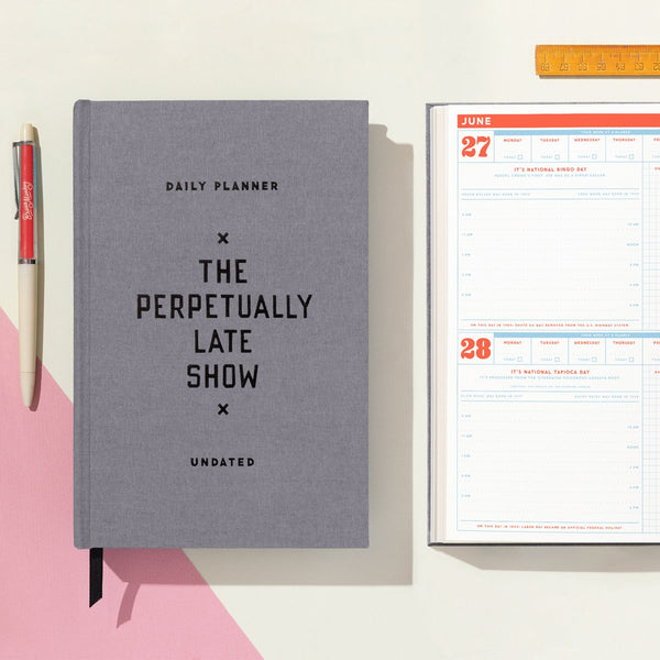 A grey cloth-bound Brass Monkey “The Perpetually Late Show Undated Standard Planner” rests beside an open daily planner displaying June 27–28, with a red pen and orange ruler on a pink and white background.