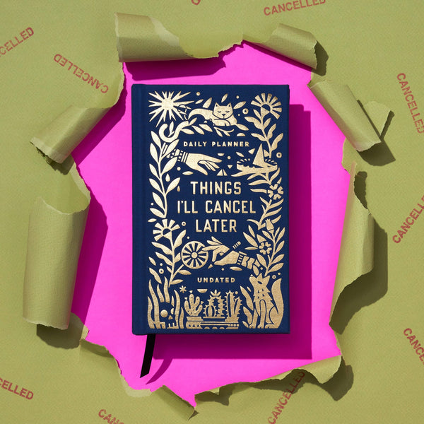 The Brass Monkey Things Ill Cancel Later Undated Mini Planner in navy blue with gold accents sits on a pink backdrop, surrounded by pieces of brown paper stamped CANCELLED.