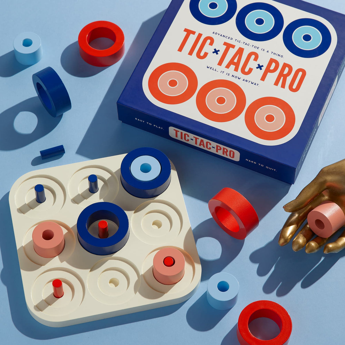 Brass Monkey’s Tic Tac Pro Game Set features painted wood blue and red pieces—circles and cylinders—on a sleek white board, with a gold hand holder, all atop a light blue surface for a modern touch.