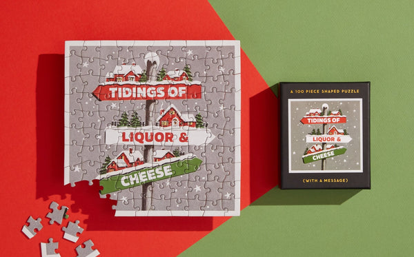 The completed Tidings 100 Piece Mini Shaped Puzzle by Brass Monkey, with its giftable box, sits on a red and green background. The design features festive houses, trees, and “Tidings of Liquor & Cheese,” with loose pieces at the bottom left corner.