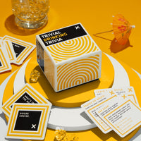 Brass Monkeys Trivial Drinking Trivia card box sits on stacked white plates, with trivia cards scattered around and a drink with ice and an umbrella nearby—all on a bright yellow background.