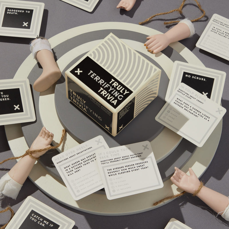 The Truly Terrifying Trivia set by Brass Monkey rests on a round platform, encircled by cards and fake severed doll arms tied with string, setting an eerie scene perfect for fans of spooky true crime trivia.