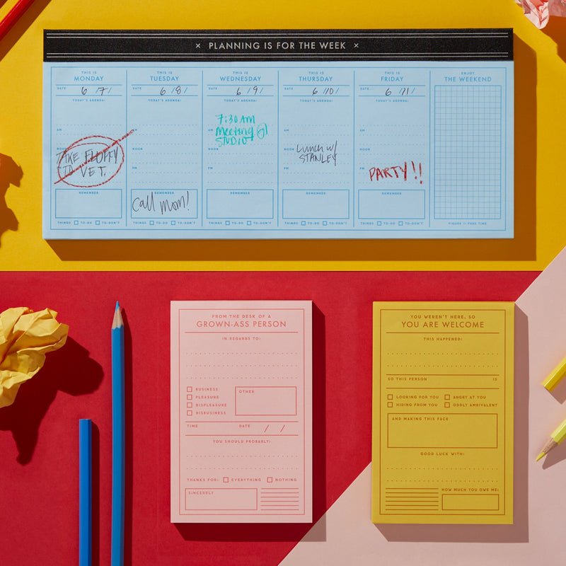 A weekly planner pad with handwritten notes on dyed paper, two pencils, and crumpled yellow paper sit on a red and yellow background. Brass Monkeys You Are Welcome Memo Pad completes the vintage-inspired arrangement.