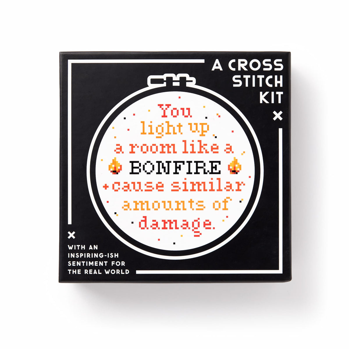A black box labeled You Light Up A Room Cross Stitch Kit by Brass Monkey displays a circular embroidery with stitched text: You light up a room like a BONFIRE + cause similar amounts of damage, surrounded by flames and decorative details.
