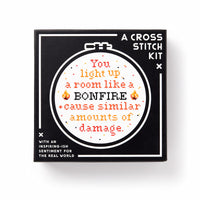 A black box labeled You Light Up A Room Cross Stitch Kit by Brass Monkey displays a circular embroidery with stitched text: You light up a room like a BONFIRE + cause similar amounts of damage, surrounded by flames and decorative details.