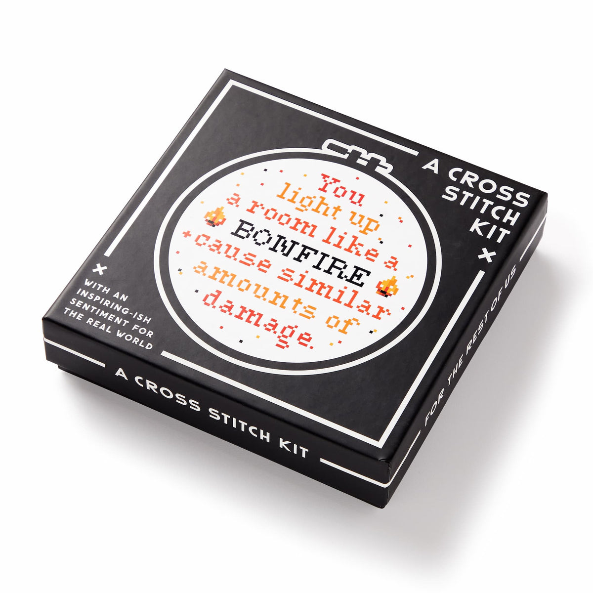 The Brass Monkey You Light Up A Room Cross Stitch Kit features a black box with a circular embroidery hoop design that reads, You light up a room like a BONFIRE & cause similar amounts of damage, accented with orange and yellow patterns.