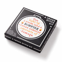 The Brass Monkey You Light Up A Room Cross Stitch Kit features a black box with a circular embroidery hoop design that reads, You light up a room like a BONFIRE & cause similar amounts of damage, accented with orange and yellow patterns.