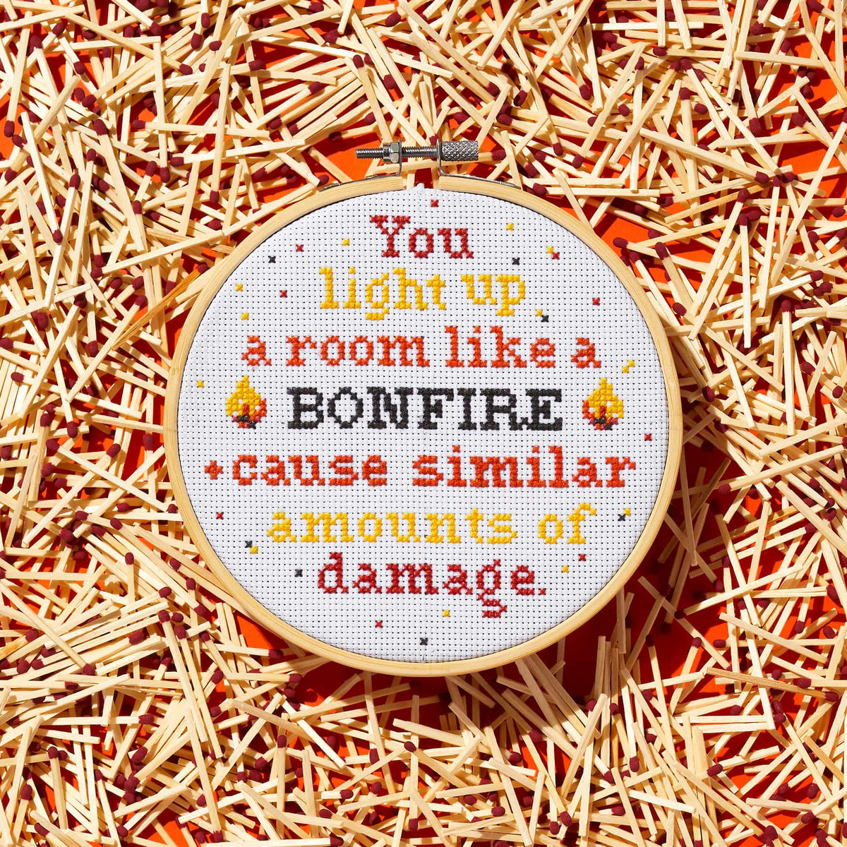 The Brass Monkey You Light Up A Room Cross Stitch Kit includes a round embroidery hoop with stitched text, match graphics on an orange background, and a design guide for easy stitching.