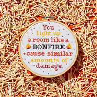 The Brass Monkey You Light Up A Room Cross Stitch Kit includes a round embroidery hoop with stitched text, match graphics on an orange background, and a design guide for easy stitching.