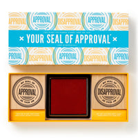 The Brass Monkey Your Seal Of Approval Stamp Set includes two oversized red stamps labeled APPROVAL and DISAPPROVAL, plus a large inkpad. The box features colorful circular designs inspired by stamped impressions.