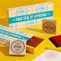The Brass Monkey Your Seal Of Approval Stamp Set is displayed on a yellow background with large red “My Seal of Approval” and “My Seal of Disapproval” stamps, a color inkpad, and a stamped sheet beside the boxed set.