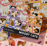 The Brass Monkey Moody Cats 1000 Piece Puzzle box displays colorful illustrations of various cat breeds with amusing captions, featuring the puzzle pieces and part of the Moody Cats puzzle below.