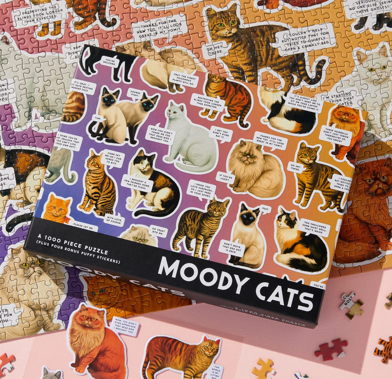 The Brass Monkey Moody Cats 1000 Piece Puzzle box displays colorful illustrations of various cat breeds with amusing captions, featuring the puzzle pieces and part of the Moody Cats puzzle below.