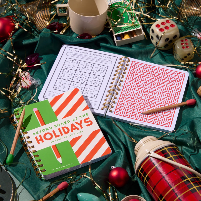 A festive flat lay featuring two open Brass Monkey puzzle books—one with sudoku, one with a red maze—surrounded by ornaments, string lights, dice, and a striped notebook titled "Beyond Bored At The Holidays.