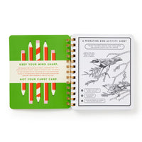 The open spiral-bound "Beyond Bored At The Holidays" notebook by Brass Monkey displays colored pencils on a green page and a black-and-white migrating bird activity sheet with illustrations and instructions on the right.