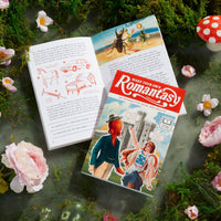 A magical scene featuring an open illustrated book and a closed Make Your Own Romantasy Book by Brass Monkey resting on moss, surrounded by flowers, mushrooms, and greenery for a whimsical, fairytale vibe.