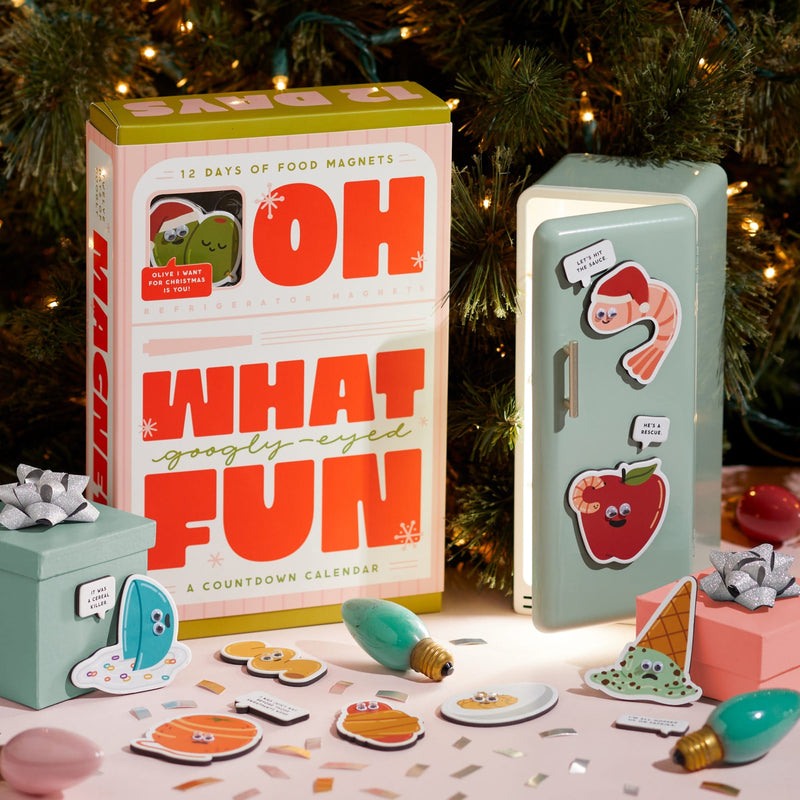 A festive scene features Brass Monkey’s Oh What Googly-Eyed Fun Magnet Countdown Calendar, a playful toy fridge with quirky food magnets, and gift boxes with bows surrounded by holiday lights and ornaments.