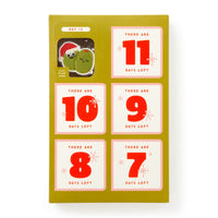 The Brass Monkey Oh What Googly-Eyed Fun Magnet Countdown Calendar features six red-labeled perforated doors counting down the days. The top left door is open, revealing a festive magnet with an avocado in a Santa hat.