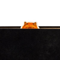 A Moody Cats Magnetic Bookmark by Brass Monkey, featuring bright orange fur and yellow eyes, peeks over a closed black book so only the cat's eyes and ears are visible.
