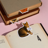 Three books feature the Brass Monkey Moody Cats Magnetic Bookmark Set. An open book shows a tabby cat bookmark with a sign reading “I heard the world sucks,” while two closed books have cat face bookmarks peeking from their pages.