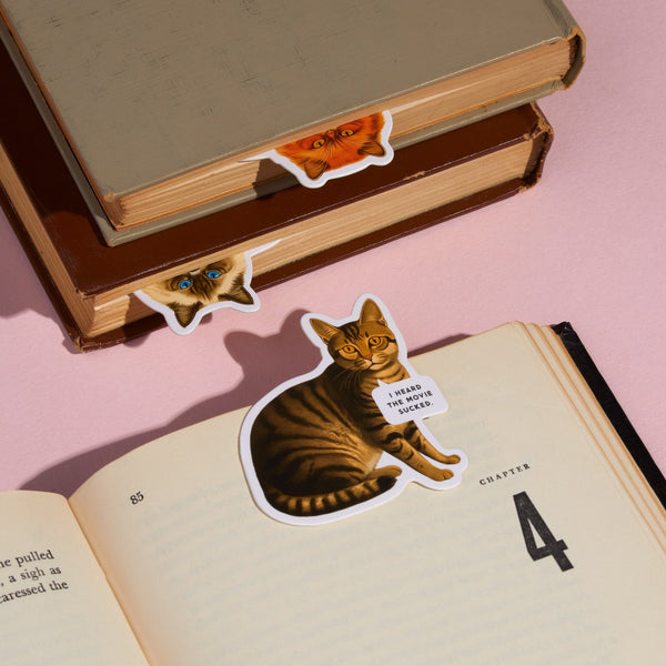 Three books feature the Brass Monkey Moody Cats Magnetic Bookmark Set. An open book shows a tabby cat bookmark with a sign reading “I heard the world sucks,” while two closed books have cat face bookmarks peeking from their pages.