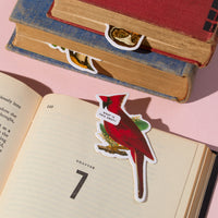 The Salty Birds Magnetic Bookmark Set by Brass Monkey is shown marking a page, featuring a red cardinal with a "What is this, 7th?" speech bubble. Two more closed books with animal bookmarks are in the background on a pink surface.