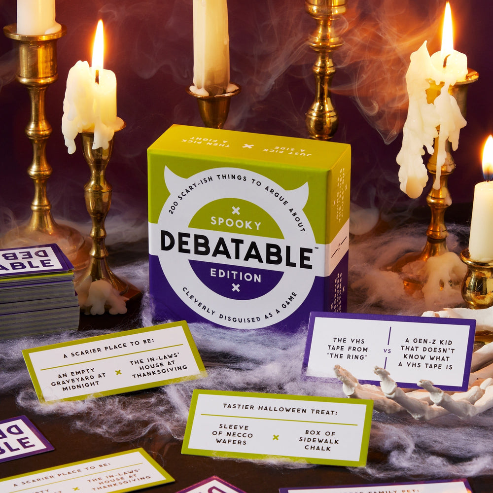 A Debatable Spooky Edition game box by Brass Monkey sits on a table with lit candles, swirling fog, and Halloween-themed debate cards, setting a mysterious and festive scene.