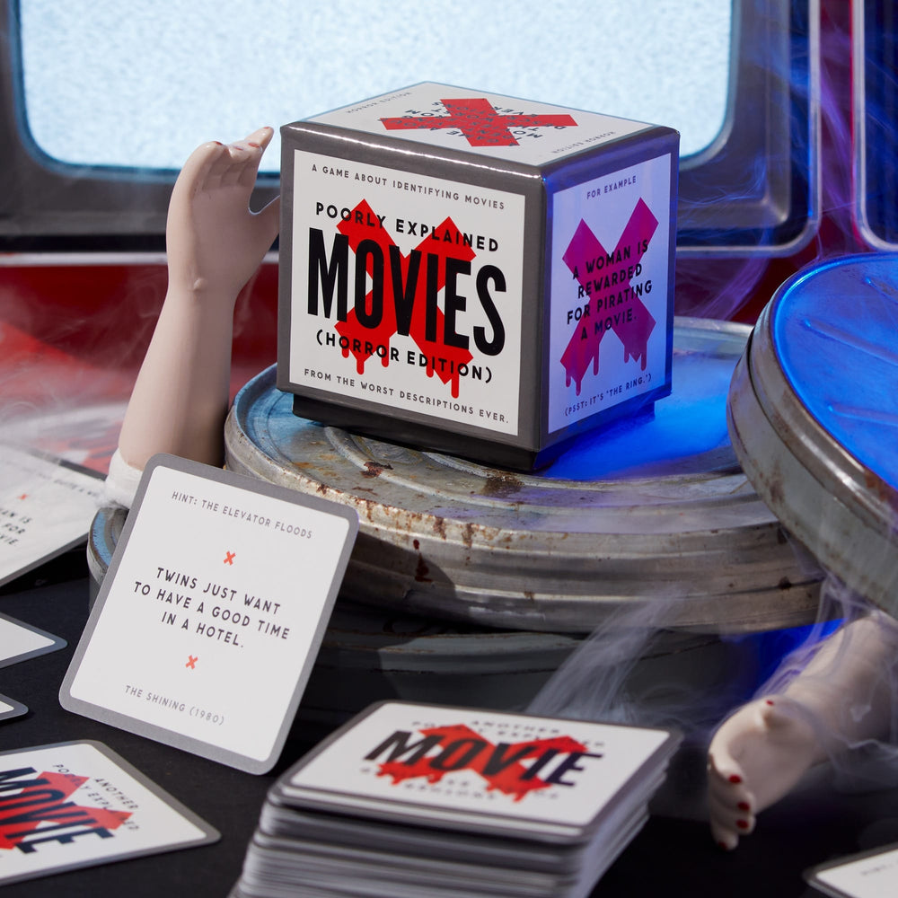A tabletop displays Brass Monkey's Mini Poorly Explained Movies - Horror Edition, featuring the game box, cards with vague horror movie descriptions, a film reel, and mannequin limbs.