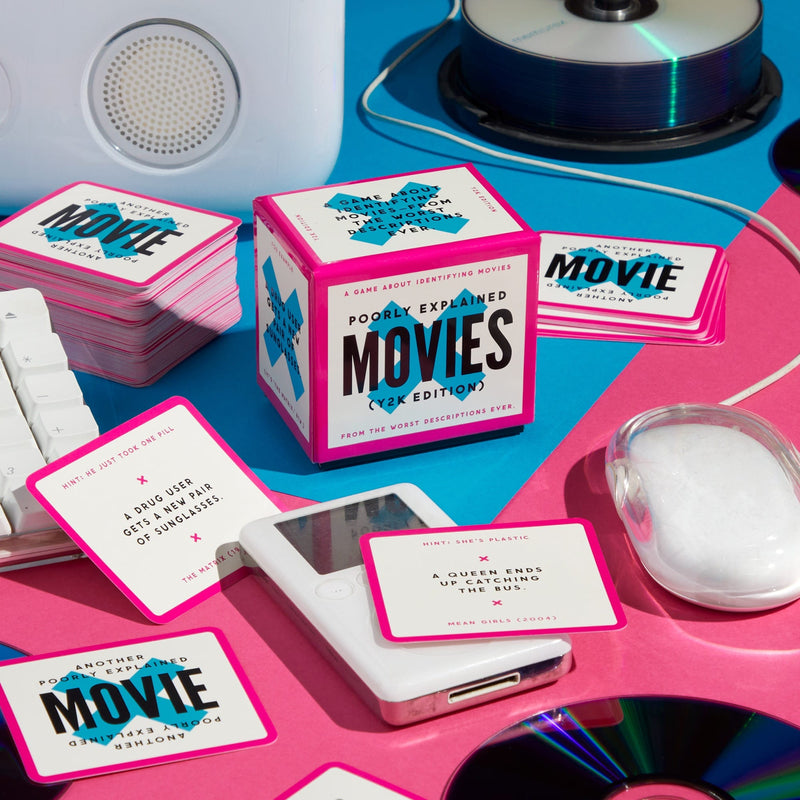 A colorful tabletop scene showcases Brass Monkey’s Mini Poorly Explained Movies – Y2K Edition, with playful movie description cards, a pink-edged box, a computer mouse, CDs, and a speaker on a blue and pink background.