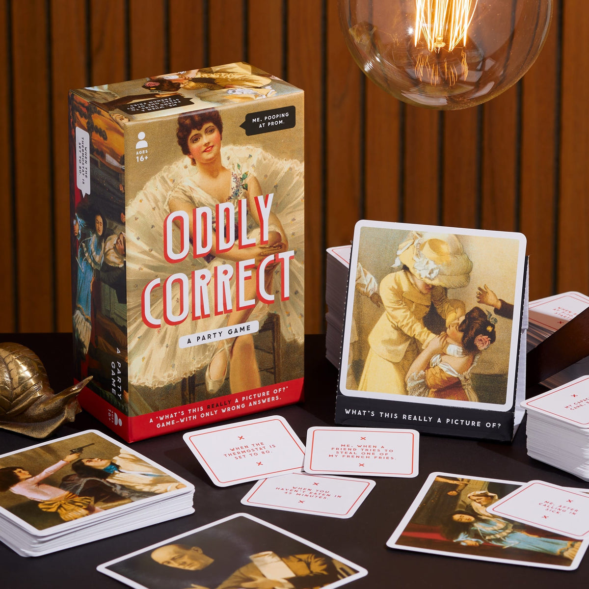 The Brass Monkey Oddly Correct Party Game is shown with its box, vintage art playing cards, and red-bordered prompt cards arranged on a table beneath a hanging light bulb.