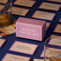 The Brass Monkey Emergency Pick-up Lines Card Set sits on a table, surrounded by scattered conversation starter cards, two drinks in glasses, a lipstick, and pills—all atop a dark blue tablecloth under warm lighting.