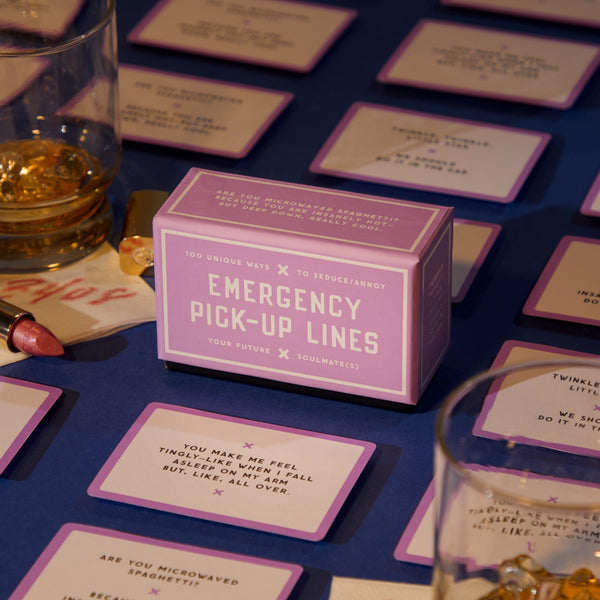 A box of Brass Monkey’s Emergency Pick-up Lines Card Set sits on a blue table, surrounded by flirty conversation starter cards, whiskey glasses, lipstick, and gold jewelry.