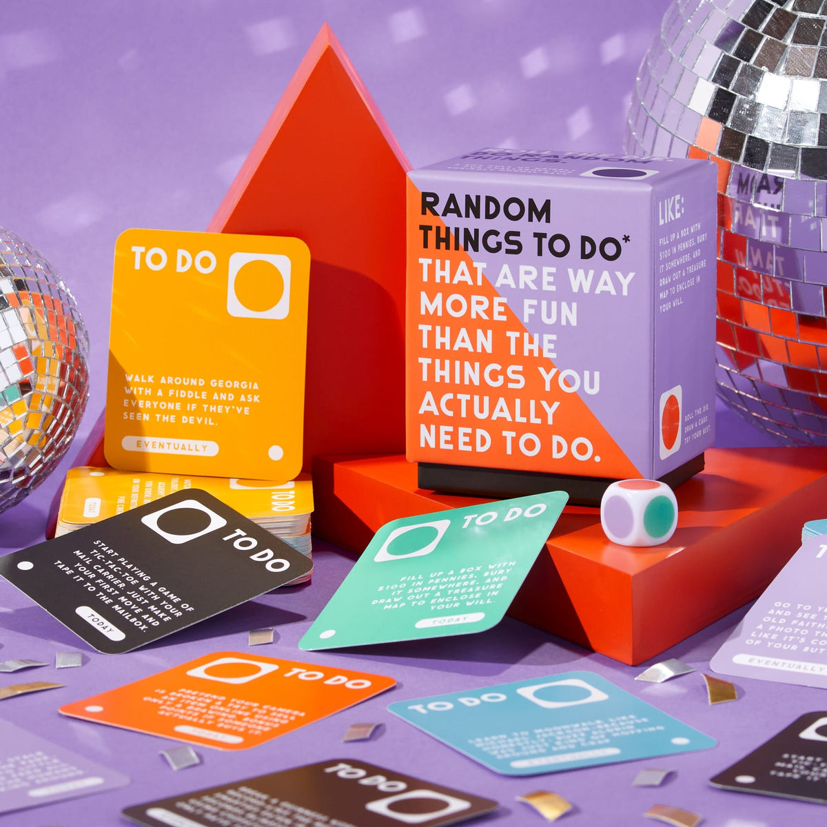 A vibrant party scene features a large dice, shiny disco balls, and bright "TO DO" cards from the Random Things To Do Box by Brass Monkey, with confetti and geometric shapes spread across a purple background.