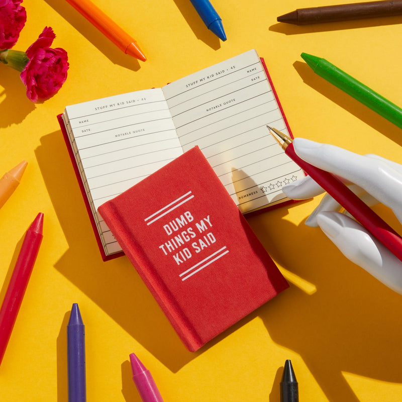 A red Brass Monkey Dumb Things My Kid Said Tiny Diary sits on a yellow surface near an open notebook, colorful markers, pink flowers, and a white-gloved hand with a red pen—perfect for capturing funny kid quotes.