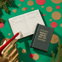 A gold-painted hand holds a red pencil above an open Brass Monkey Naughty and/or Nice Tiny Diary on a green surface with gold, red, and brown circles, accented by pine branches in the corners.