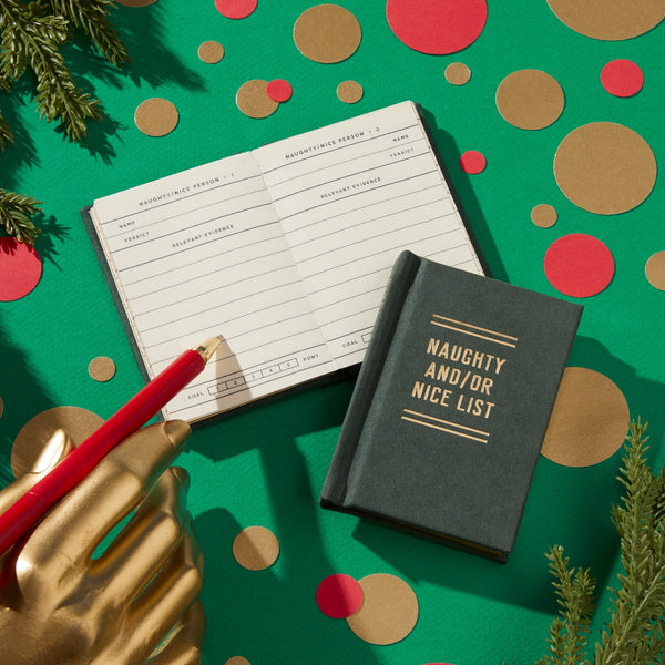 A gold-painted hand holds a red pencil above an open Brass Monkey Naughty and/or Nice Tiny Diary on a green surface with gold, red, and brown circles, accented by pine branches in the corners.