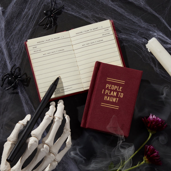 A red People I Plan To Haunt Tiny Diary by Brass Monkey lies open on a table with cobwebs, black spiders, a white candle, a skeleton hand holding a pen, and purple flowers for a spooky Halloween scene.