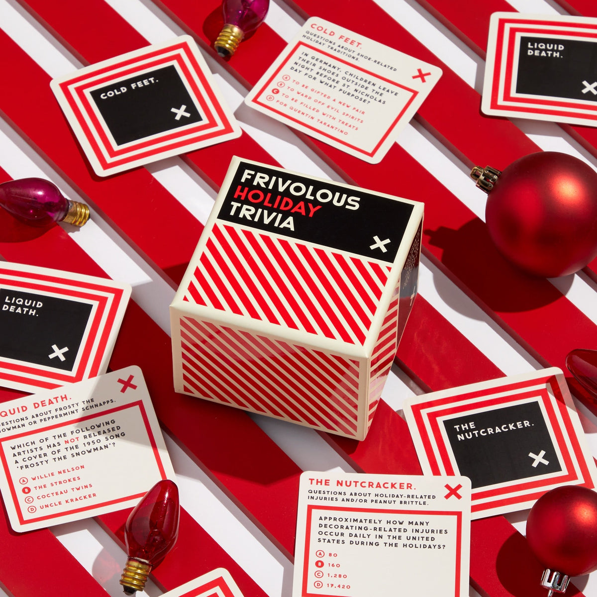 Brass Monkey’s Frivolous Holiday Trivia game sits among trivia cards, red ornaments, and Christmas lights on a striped surface. The cards feature multiple-choice holiday questions.