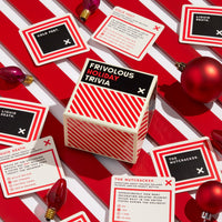 Brass Monkey’s Frivolous Holiday Trivia game sits among trivia cards, red ornaments, and Christmas lights on a striped surface. The cards feature multiple-choice holiday questions.