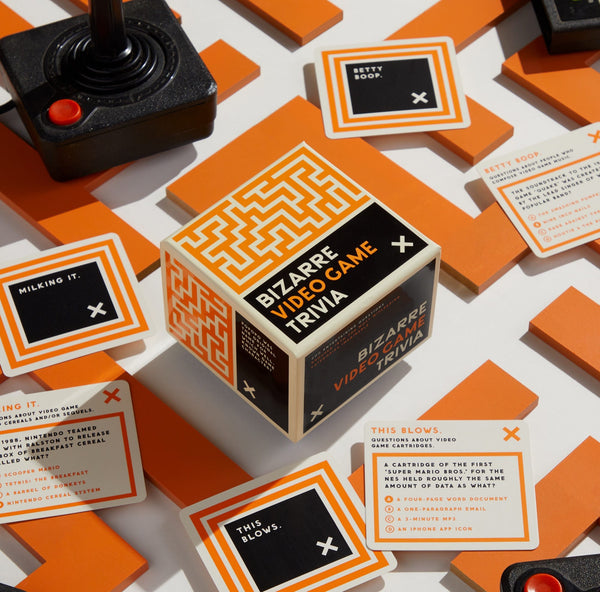 A tabletop display features a Brass Monkey Bizarre Video Game Trivia box in orange and black, vintage joystick, trivia cards, and orange tiles, all arranged on a white background.