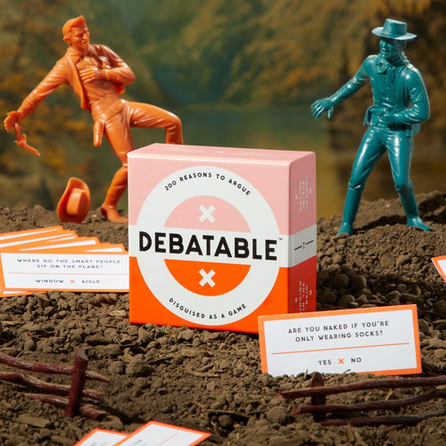 A box of the Debatable Game Set by Brass Monkey rests on dirt among plastic cowboy figures and quirky question cards—making it an ideal social game for your next Brass Monkey gathering.