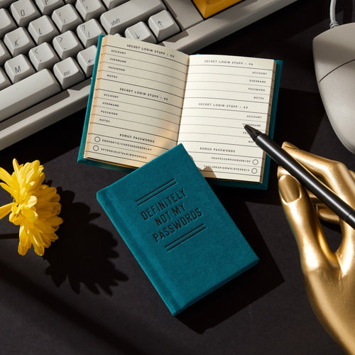 The Brass Monkey “Definitely Not My Passwords” Password Diary in teal sits open on a desk with a keyboard, yellow flower, and gold hand-shaped pen holder with a black pen, sunlight casting shadows across the scene.