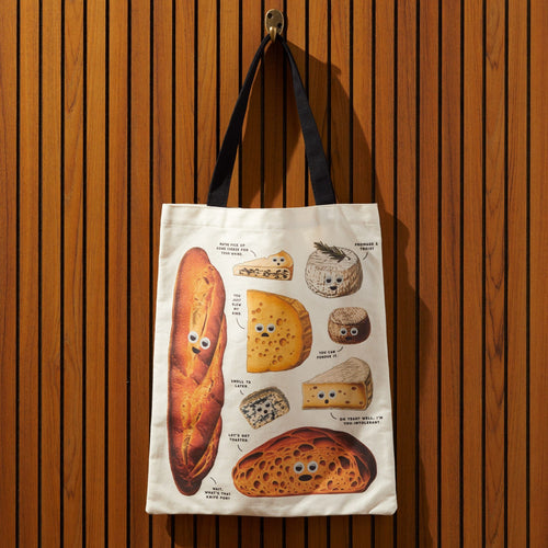 The Brass Monkey Googly Food Canvas Tote, featuring playful breads and cheeses with googly eyes, hangs on a wooden hook before vertical slats—a quirky foodie gift.