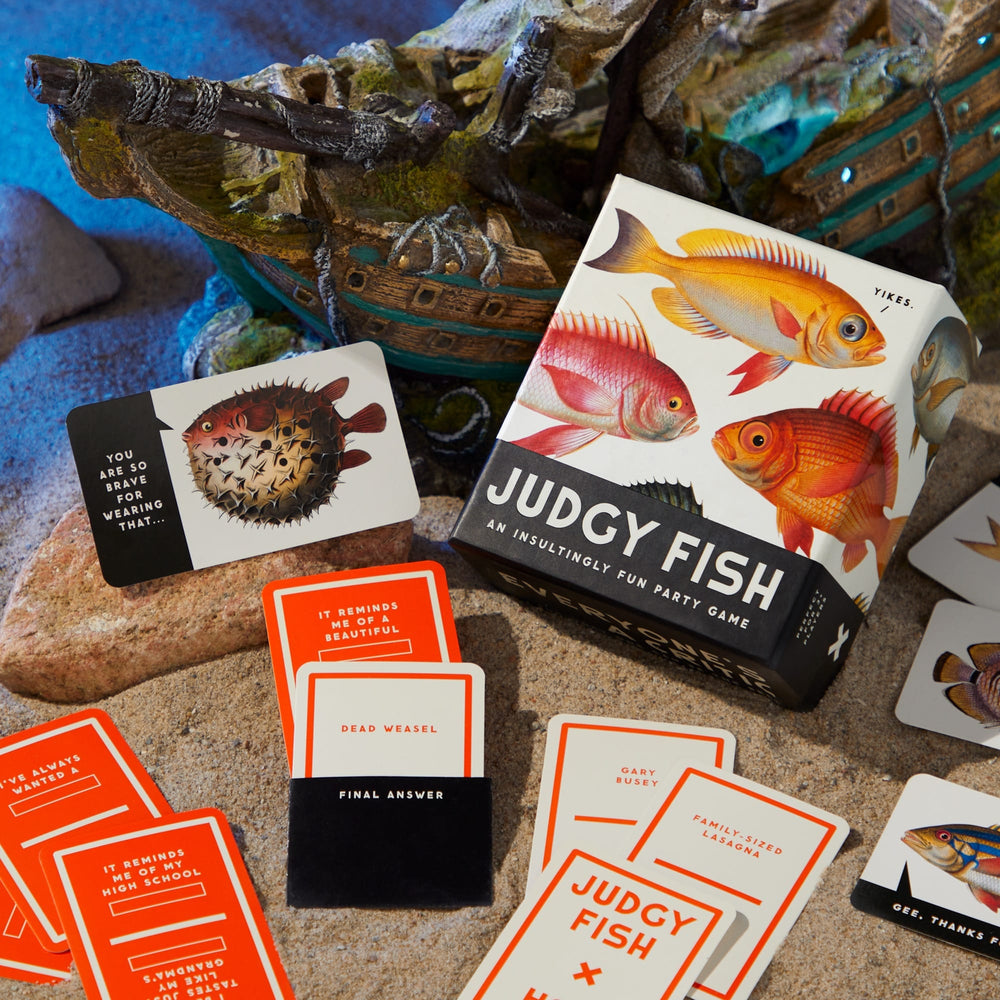 The Brass Monkey Judgy Fish Game is a colorful adult party set, with cards spread on sand, a box showing illustrated fish, and a decorative ship nearby. Some cards feature funny phrases for extra playful fun.