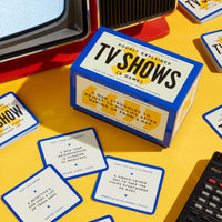 A blue-and-yellow box labeled "Poorly Explained TV Shows Game" by Brass Monkey sits on a yellow surface with clue cards featuring funny pop culture references and retro items—perfect for your next party game night.